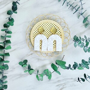 Puede incluir: Pendientes blancos en forma de arco con un patrón de helecho, presentados sobre una base circular tejida. Los pendientes tienen tachuelas doradas en forma de diamante. Hojas de eucalipto dispuestas sobre una superficie de mármol.