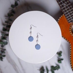 Puede incluir: Pendientes colgantes con un disco azul claro y perlas blancas. Los pendientes cuelgan de ganchos plateados. Se muestran sobre una superficie blanca. En el fondo, un ukelele.