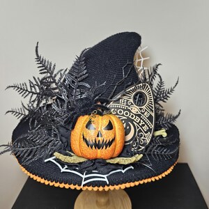 May include: A black witch hat decorated with a carved orange pumpkin, black fern accents, and a decorative Ouija board. The hat has an orange trim and a white spiderweb design.