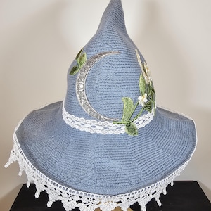 May include: A blue pointed hat featuring a silver crescent moon emblem, green embroidered leaves, and small white flowers. The hat is decorated with a white lace band around the base and a wide white lace trim along the edge of the brim.