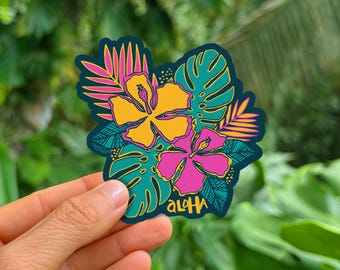 May include: A die-cut sticker featuring a vibrant illustration of two hibiscus flowers in yellow and pink, surrounded by teal and pink tropical palm and monstera leaves. The word Aloha is printed in a stylized yellow font at the bottom center.