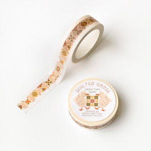 Puede incluir: Un rollo de cinta washi con un patrón de cuadrados de patchwork en tonos naranja, marrón y blanco. La cinta está etiquetada como "Quilted Goose Washi Tape 15mm" y "Diseñado por Anna Parttinen annaparttinen.com".