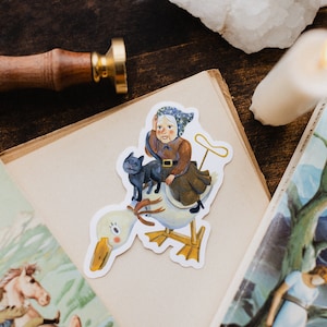 Op de afbeelding: Een grillige sticker met een vrouw die op een gans rijdt, vergezeld van een zwarte kat. De vrouw draagt een bruine jurk en een blauwe hoofddoek. De gans is wit met gele poten en een lange oranje snavel. De sticker heeft een vintage illustratiestijl.