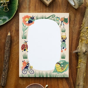 May include: A decorative notepad with a whimsical illustration of anthropomorphic insects. The border features a variety of bugs engaged in activities like carrying baskets and riding bicycles, set against a beige background with a white oval space for writing.