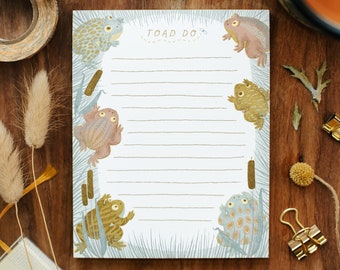 May include: A white notepad with blue and green borders features a whimsical illustration of toads and reeds. The notepad has lined pages and the text "TOAD DO" at the top.