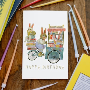 May include: A white card featuring a colourful illustration of two bunnies riding a bicycle with a book cart. The cart is filled with books and has a sign that says "Used & Local Book Bike". The card reads "Happy Birthday".