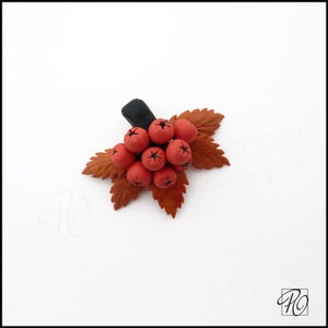 May include: A handmade polymer clay brooch featuring a cluster of red berries with black centers, set against a backdrop of brown leaves.