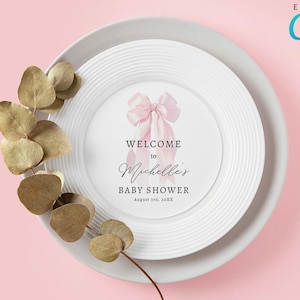 Puede incluir: Un plato blanco con un lazo rosa y el texto "Bienvenido al Baby Shower de Michelle el 17 de agosto de 20XX" sobre un fondo rosa. Editable con Canva.