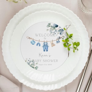 Puede incluir: Un plato blanco con una invitación de baby shower azul y verde. La invitación dice "Bienvenido a la baby shower de Kara" con una fecha y una línea de ropa colgada en un tendedero. La invitación es editable con Canva.