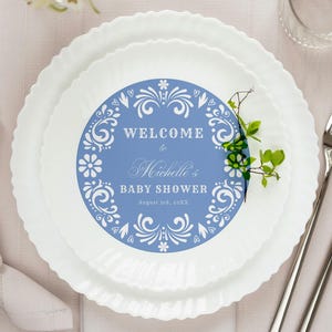 Puede incluir: Una mesa para baby shower con platos blancos, un plato decorativo azul y blanco, cubiertos y una servilleta doblada. El plato decorativo tiene el texto "WELCOME to Michelle's BABY SHOWER August 20th, 2022".