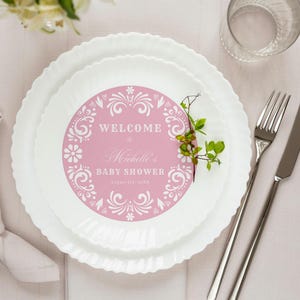 Puede incluir: Decoración de mesa para un baby shower. Un plato decorativo rosa y blanco que dice "Welcome to Michelle's Baby Shower". El plato está rodeado de platos blancos, cubiertos, una servilleta y un vaso.