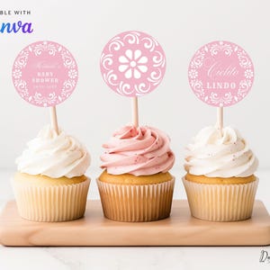 Puede incluir: Tres cupcakes con glaseado rosa y blanco, cubiertos con adornos decorativos rosas y blancos sobre palitos de madera. Los adornos tienen diseños florales y de texto. Los cupcakes están sobre una tabla de madera. El texto de la imagen dice "Editable with Canva".