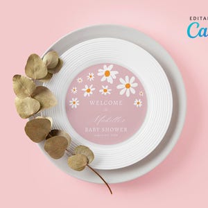 Puede incluir: Un plato blanco con un círculo rosa en el centro. El círculo tiene un patrón de margaritas blancas y el texto "Bienvenido al Baby Shower de Michelle" en blanco. El plato está sobre un fondo rosa con una rama de hojas de eucalipto secas.