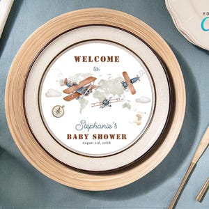 Puede incluir: Un diseño de plato para baby shower imprimible con una ilustración de acuarela de dos aviones volando sobre un mapa del mundo. El texto dice "Bienvenido a la fiesta de bienvenida al bebé de Stephanie, 3 de agosto de 20XX".