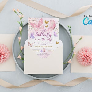May include: A baby shower invitation with a butterfly theme. The invitation features pink and purple butterflies and the text "a little Butterfly is on the way!". The invitation is inside a cream-colored envelope, with pink floral accents.