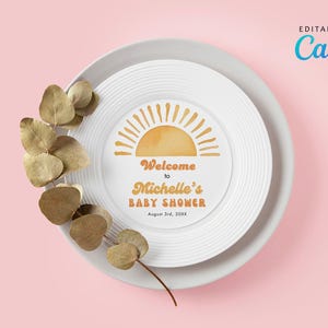 Puede incluir: Un plato blanco con un gráfico redondo de sol naranja y amarillo con el texto "Bienvenido al Baby Shower de Michelle el 3 de agosto de 20XX". El plato está sobre un fondo rosa con una rama de eucalipto seco.