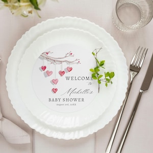 Puede incluir: Un plato blanco con un diseño de acuarela rosa y blanco de una rama con corazones colgando. El texto "Bienvenido al Baby Shower de Michelle" está impreso en el plato. El plato está sobre un mantel rosa claro con una servilleta blanca y cubiertos.