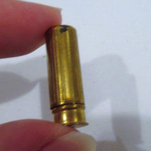May include: A cylindrical brass-coloured metal cartridge casing with a smooth, polished surface. The object features a distinct rimmed base and a hollow opening at the top, showing a metallic sheen and a small dark mark on the upper side.
