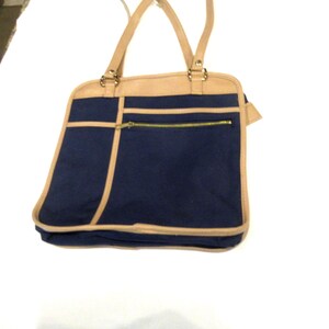 Può includere: Una borsa tote in tela blu con finiture in pelle marrone e una tasca con cerniera. La borsa ha due manici superiori.