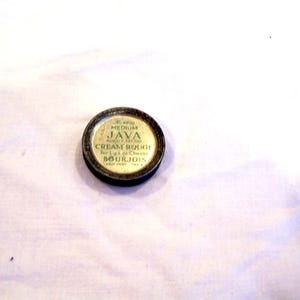 May include: A vintage round makeup compact with a black rim and a gold-colored top. The top features the text "JAVA CREAM ROUGE" and "BOURJOIS" in a classic font. The compact is for lips or cheeks.