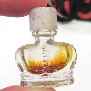 May include: A miniature clear glass perfume bottle shaped like a crown with a translucent white stopper containing gold flecks. The bottle features a textured base and a central vertical row of raised dots, containing a small amount of amber liquid.