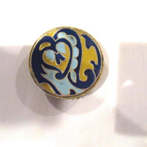 May include: A round, vintage brooch with a gold-colored metal frame. The brooch features a blue, yellow, and light blue enamel design. The design is a stylized floral or abstract pattern. The brooch is set against a white background.