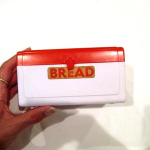 Op de afbeelding: Een witte en rode plastic brooddoos met het woord "BREAD" in een goudkleurig lettertype. De doos heeft een rood deksel en een sluiting. De brooddoos is rechthoekig en lijkt vintage.