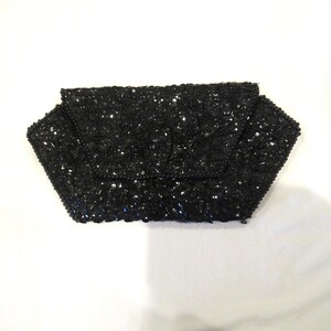 May include: Black beaded clutch with a flap closure. The clutch is covered in small, shiny black beads, giving it a textured appearance. The flap has a decorative beaded design. The clutch is on a white background.