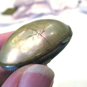 May include: An antique, oval-shaped, brass-toned container with a cracked, pearlescent surface. The container has a hinged lid and a decorative, ridged edge. The object is held in a person's hand.