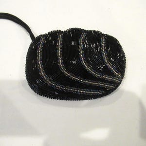 Può includere: Pochette nera con perline, dalla forma curva e con cinturino nero. La pochette è ricoperta da piccole perline nere con accenti iridescenti, creando un effetto scintillante. Il design presenta linee curve di perline.