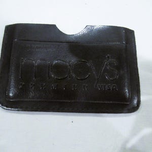 Op de afbeelding: Een donkerbruine leren kaarthouder met het Macy's Premier Visa-logo in reliëf. De kaarthouder heeft één vak en een gebogen uitsparing aan de bovenkant. Het leer lijkt glad en licht glanzend.