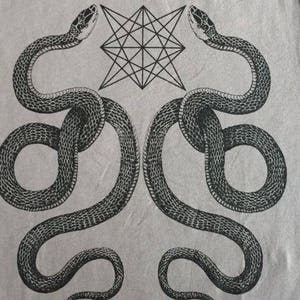 K&ouml;nnte beinhalten: Ein graues T-Shirt mit einem schwarz-wei&szlig;en grafischen Design aus zwei Schlangen, die sich um einen geometrischen Stern winden.
