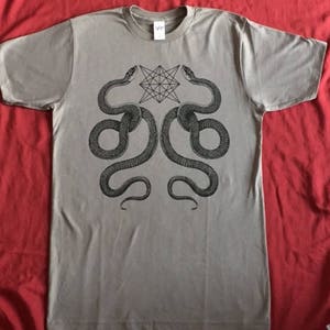 K&ouml;nnte beinhalten: Ein graues T-Shirt mit einem schwarz-wei&szlig;en Grafikdruck von zwei Schlangen, die sich gegen&uuml;berstehen, und einem geometrischen Stern in der Mitte.