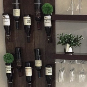 May include: A dark wood wine rack with eight bottles of wine hanging from metal holders. The rack is mounted on a wall with a shelf above it. There are four wine glasses hanging from the shelf.