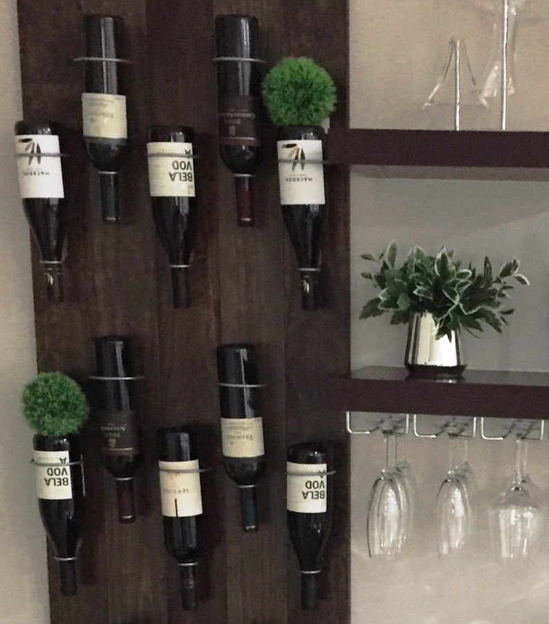 May include: A dark wood wine rack with eight bottles of wine hanging from metal holders. The rack is mounted on a wall with a shelf above it. There are four wine glasses hanging from the shelf.