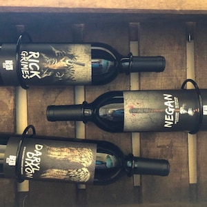 May include: Three wine bottles with labels featuring characters from the TV show The Walking Dead. The labels feature Rick Grimes, Negan, and Daryl Dixon. The bottles are displayed in a wooden wine rack.