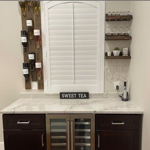 May include: A home bar with a wine rack, wine glasses, and a wine cooler. The bar has a white countertop and dark brown cabinets. The wine rack is made of wood and has a rustic look. The wine glasses are hanging from a wooden shelf. The wine cooler is stainless steel and has a glass door. The countertop has a sign that says "Sweet Tea".