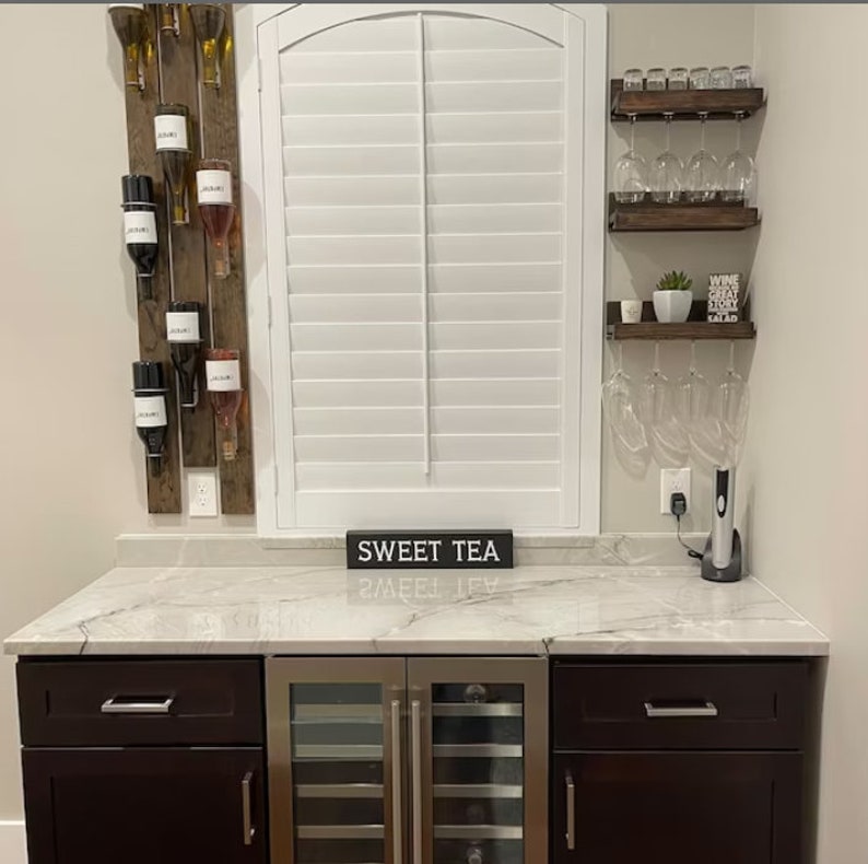 May include: A home bar with a wine rack, wine glasses, and a wine cooler. The bar has a white countertop and dark brown cabinets. The wine rack is made of wood and has a rustic look. The wine glasses are hanging from a wooden shelf. The wine cooler is stainless steel and has a glass door. The countertop has a sign that says "Sweet Tea".