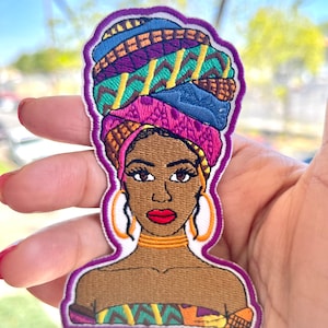 Peut inclure: Écusson brodé représentant une femme à la peau brune portant un foulard coloré, des créoles dorées et un collier doré. L'écusson a une bordure violette et est tenu dans une main.