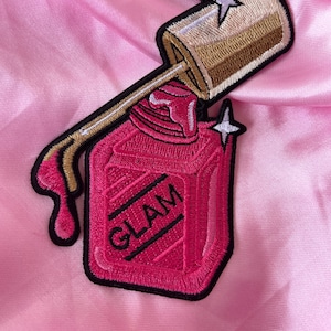 May include: Pink nail polish bottle with a gold cap and a white star on top. The bottle is embroidered with the word "GLAM" in black.