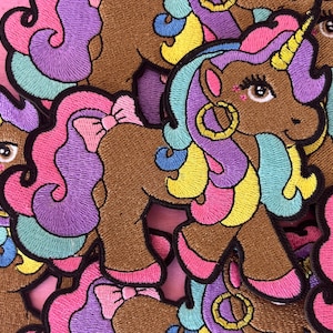 May include: Embroidered patches featuring a brown unicorn with a golden horn and hoop earrings. The unicorn has a colorful mane in shades of purple, pink, yellow, and teal. The patches are detailed with embroidered outlines and accents.