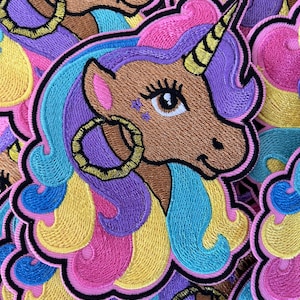 May include: A stack of colorful embroidered iron-on patches featuring a unicorn with a gold horn and a gold hoop earring. The unicorn has brown fur, pink, yellow, blue, and purple hair, and a black outline.