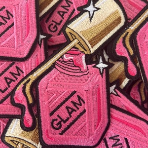 May include: Close-up of several pink embroidered iron-on patches with the word "GLAM" in black lettering. The patches are shaped like nail polish bottles with a gold cap and a pink drip.