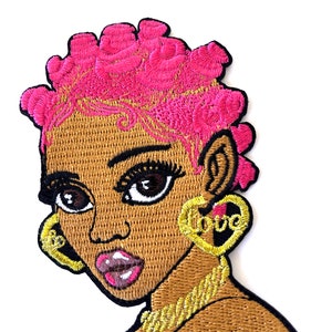May include: Embroidered iron-on patch featuring a cartoon illustration of a woman with pink hair, brown skin, and gold jewelry. The woman is wearing large hoop earrings with the word "love" on one earring.