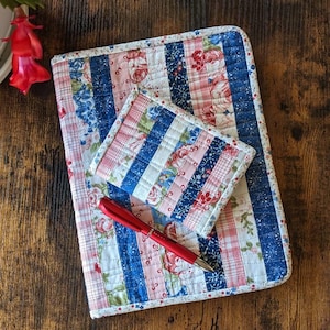May include: A quilted fabric notebook cover and a matching smaller wallet featuring vertical stripes of floral, plaid, and solid blue patterns in red, white, and blue. A red ballpoint pen rests diagonally across the center of the items.