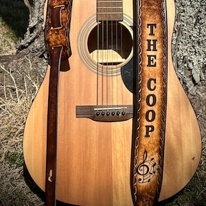 May include: A brown leather guitar strap with a decorative border and the text "THE COOP" and a musical note design. The strap is attached to an acoustic guitar.