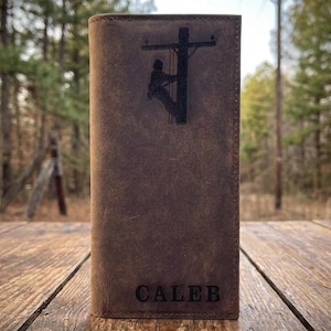 May include: A brown leather wallet with a silhouette of a lineman on a utility pole. The name "CALEB" is embossed at the bottom. The wallet is rectangular and appears to be made of distressed leather.