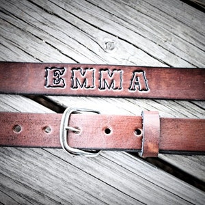 May include: A brown leather belt with a silver buckle. The belt is personalized with the name "EMMA" embossed in the leather.