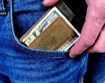 May include: A brown leather wallet with a money clip, holding a one hundred dollar bill, is partially visible in the front pocket of blue jeans. The wallet has a worn, textured appearance.