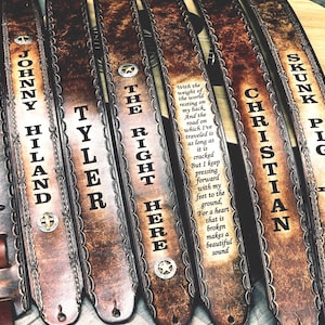 May include: A collection of brown leather guitar straps with various names and phrases embossed in white. The straps have decorative edges and metal accents. One strap features a quote, and another has a star.